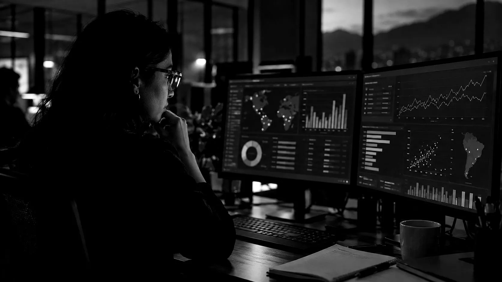 Una persona de perfil frente a dos monitores con gráficas de datos, en una oficina en penumbra.